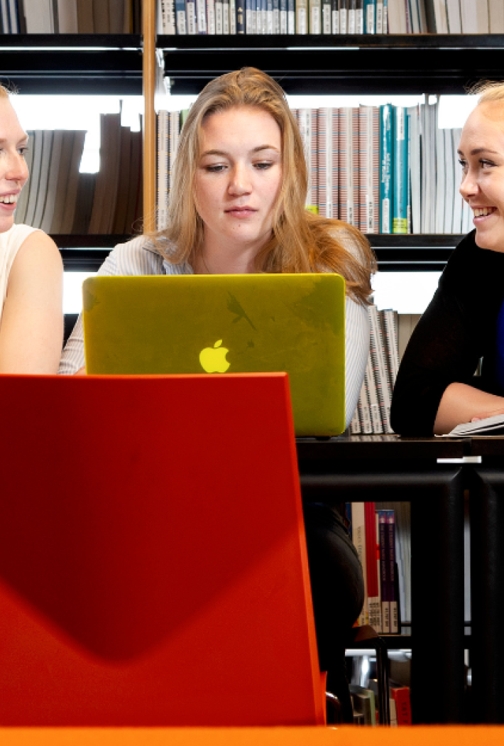 Tre unge kvinder sidder ved et bord i et bibliotek og arbejder sammen ved en bærbar computer, omgivet af åbne bøger og reoler fyldt med materialer i baggrunden.        Spørg ChatGPT